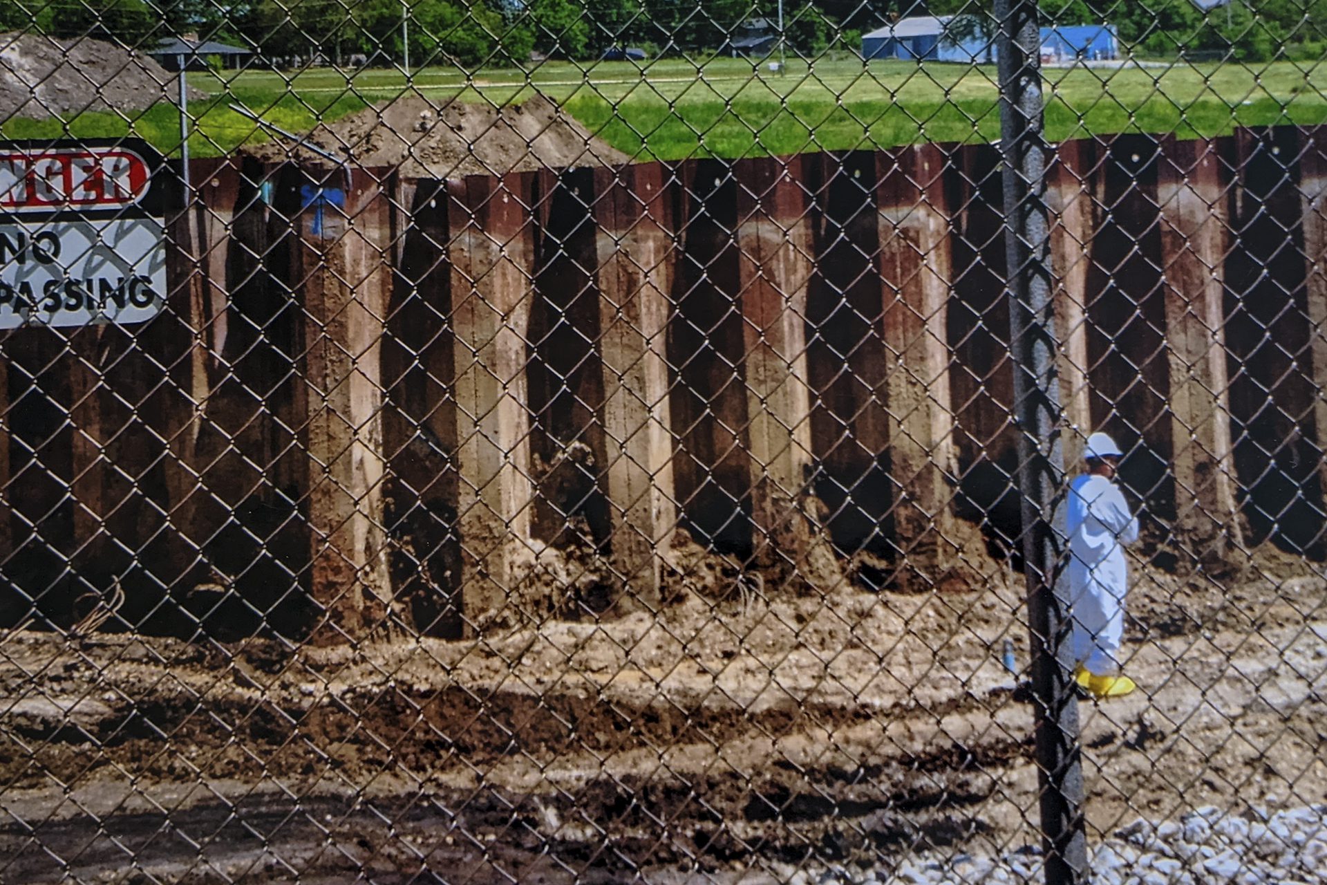 A person in full body white suit helmet and yellow boots in a pit behind a fence with a sign that reads "danger, no trespassing"