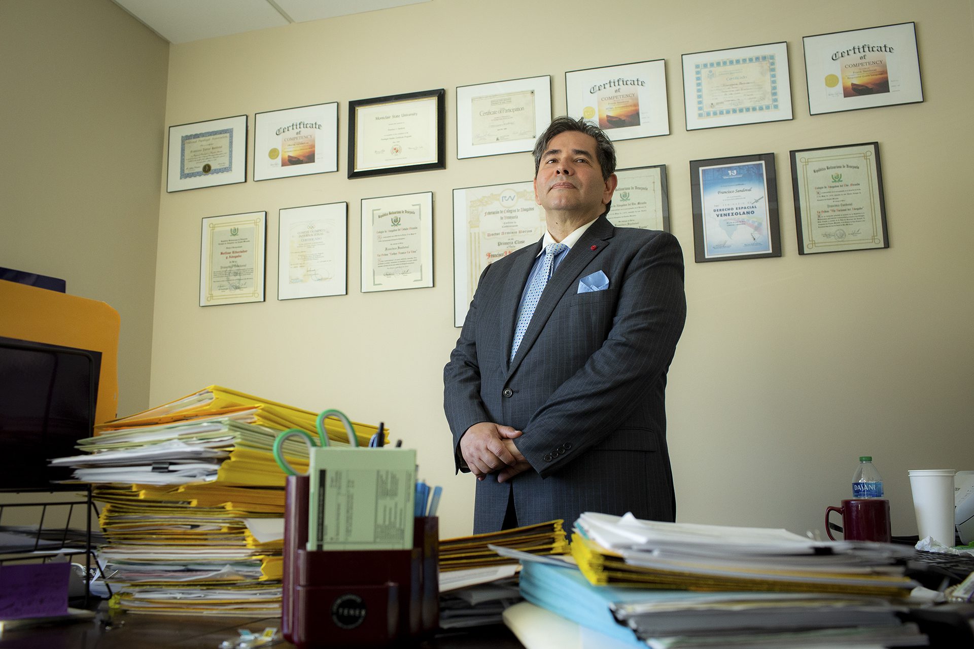 Frank Sandoval wearing a gray jacket and blue dot tie in front of various framed awards and certificates in his office