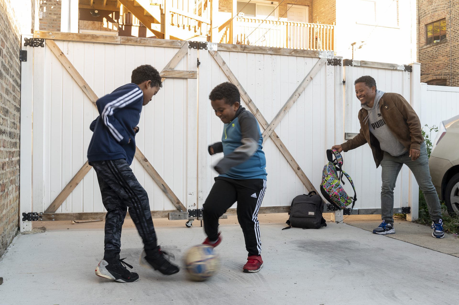 Two children play soccer as a man smiles while holding a backpack.