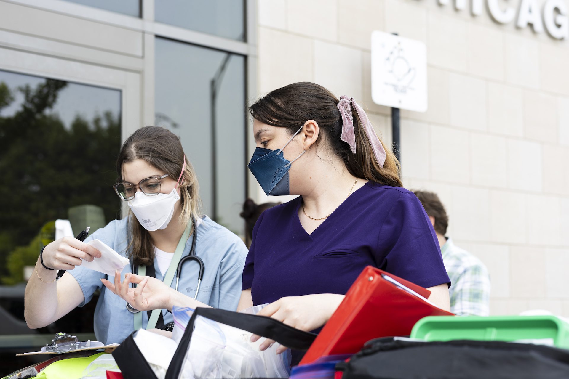 Dos estudiantes de medicina con bata y mascarilla examinan material médico en el exterior de una comisaría.