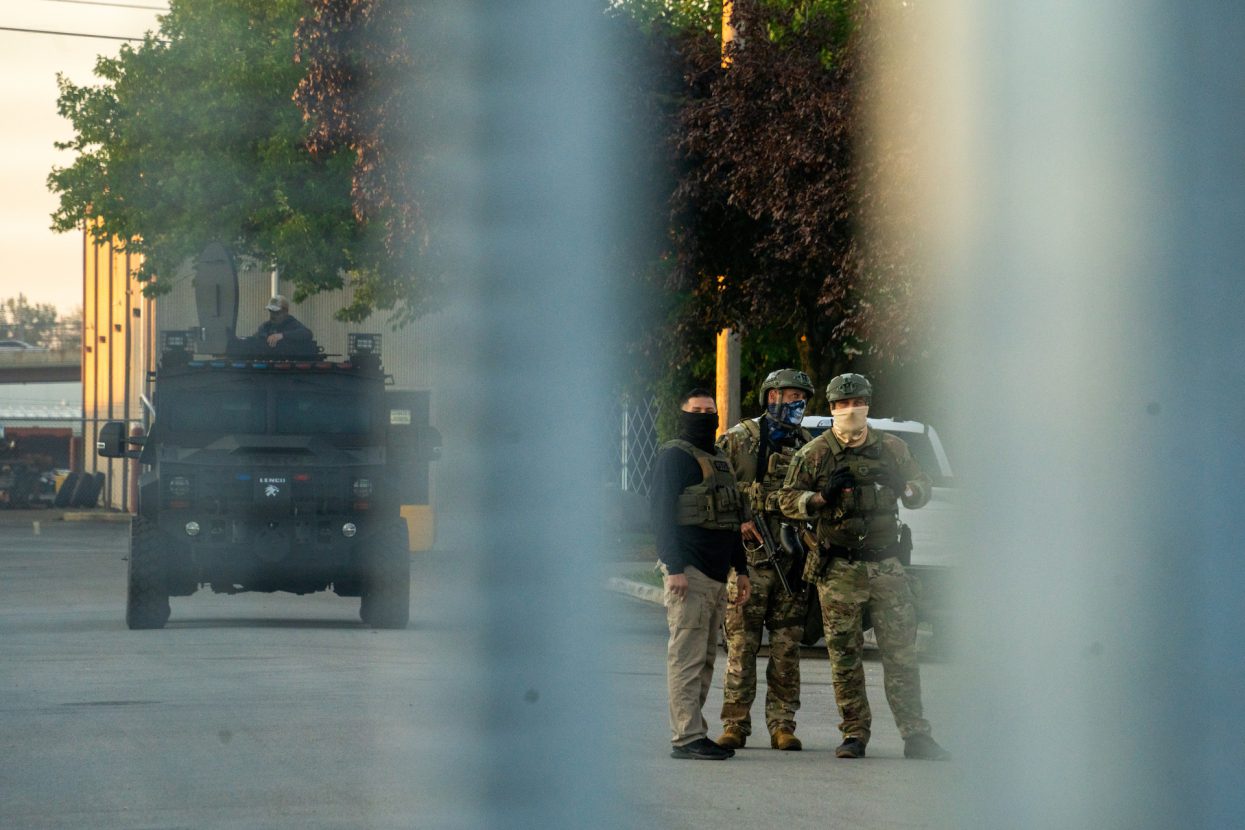 Three masked federal agents stand behind a perimeter fence at a detenion center.