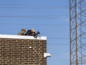 Two federal agents wearing camouflage and standing on a rooftop aim less lethal weapons at protesters.