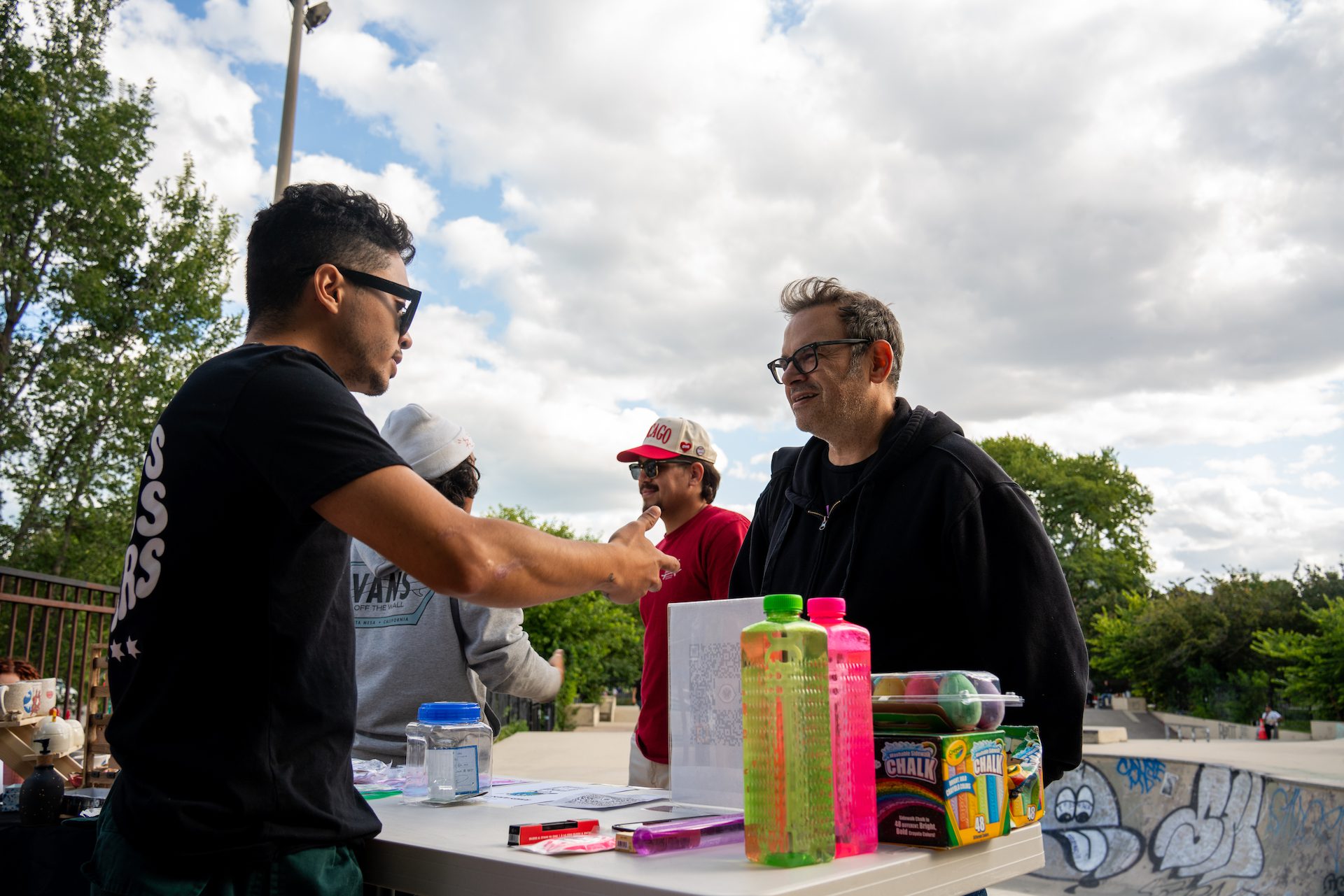 Javier Ponce stands behind a table as he explains the mission of Boards across Borders to a man wearing glasses during a fundraising event in partnership with Chicago Roller Girls at Wilson Skate Park on
