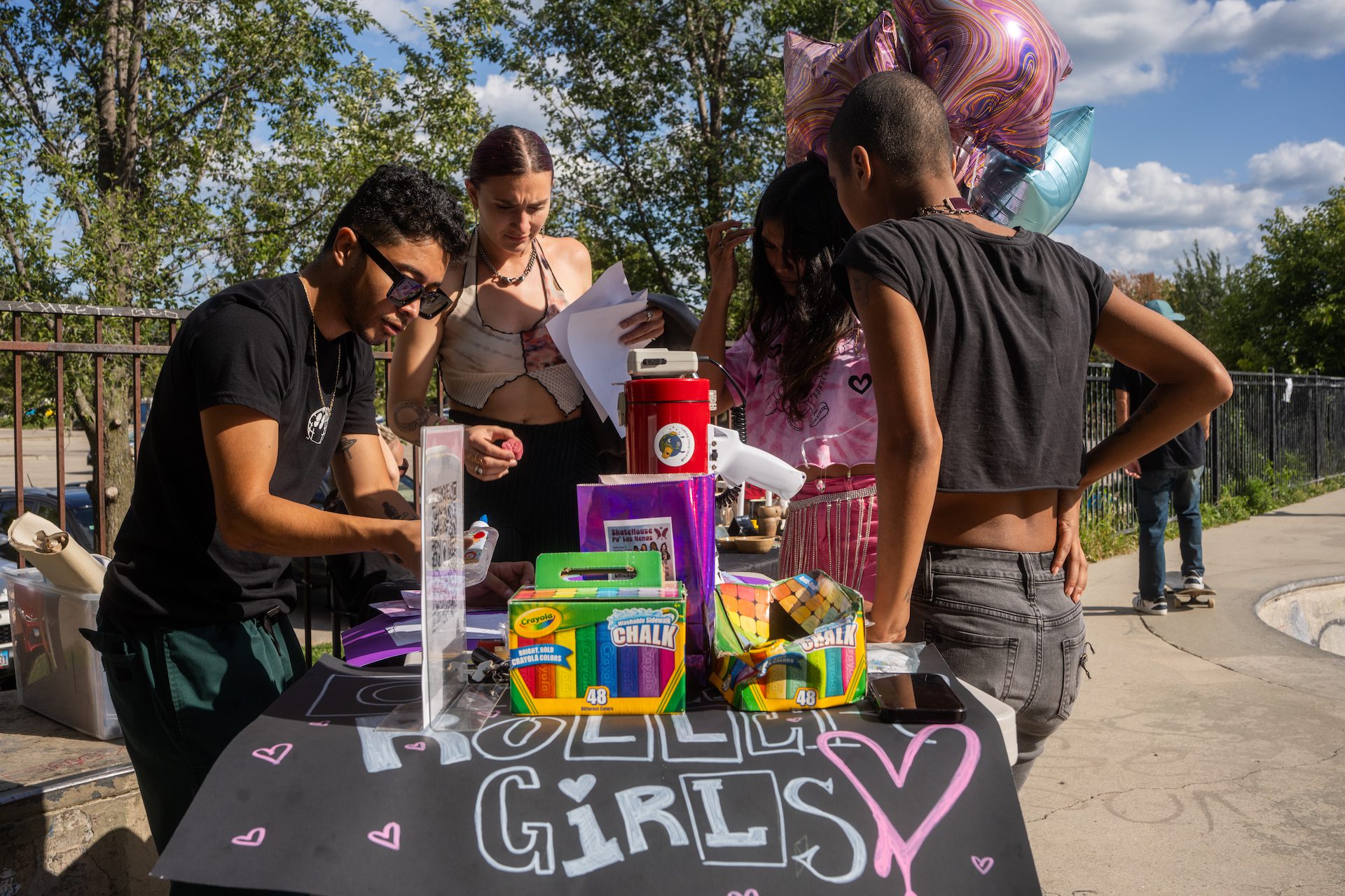 Javier Ponce stands behind a table with colorful chalk as he assembles a poster during a Boards Across Borders fundraising event in partnership with Chicago Roller Girls at Wilson Skate Park on a sunny Saturday.