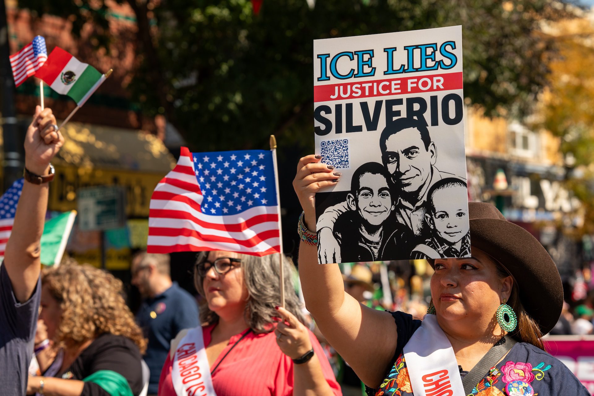 A participant in the Little Village Mexican Independence Day parade wearing a cowboy hat carries a sign in support of Silverio Villegas-Gonzalez. People around her carry Mexican and American flags.