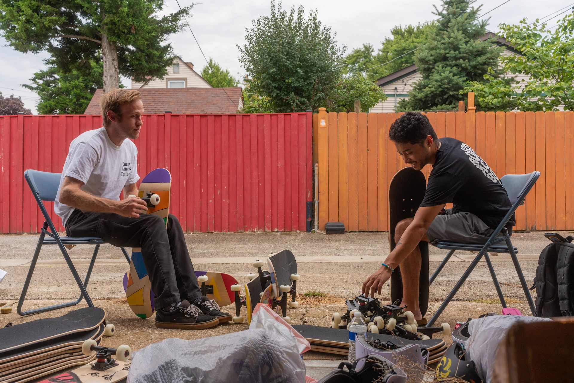 Friends Javier Ponce and C.DOT sit in an open garage on folding chairs disassembling donated skate boards to make them easier to pack during the boxing process.