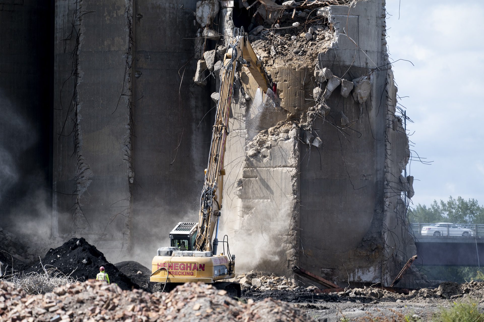 A construction crane tears down concrete from the remaining silo tower. Rubble surrounds the demolition site.