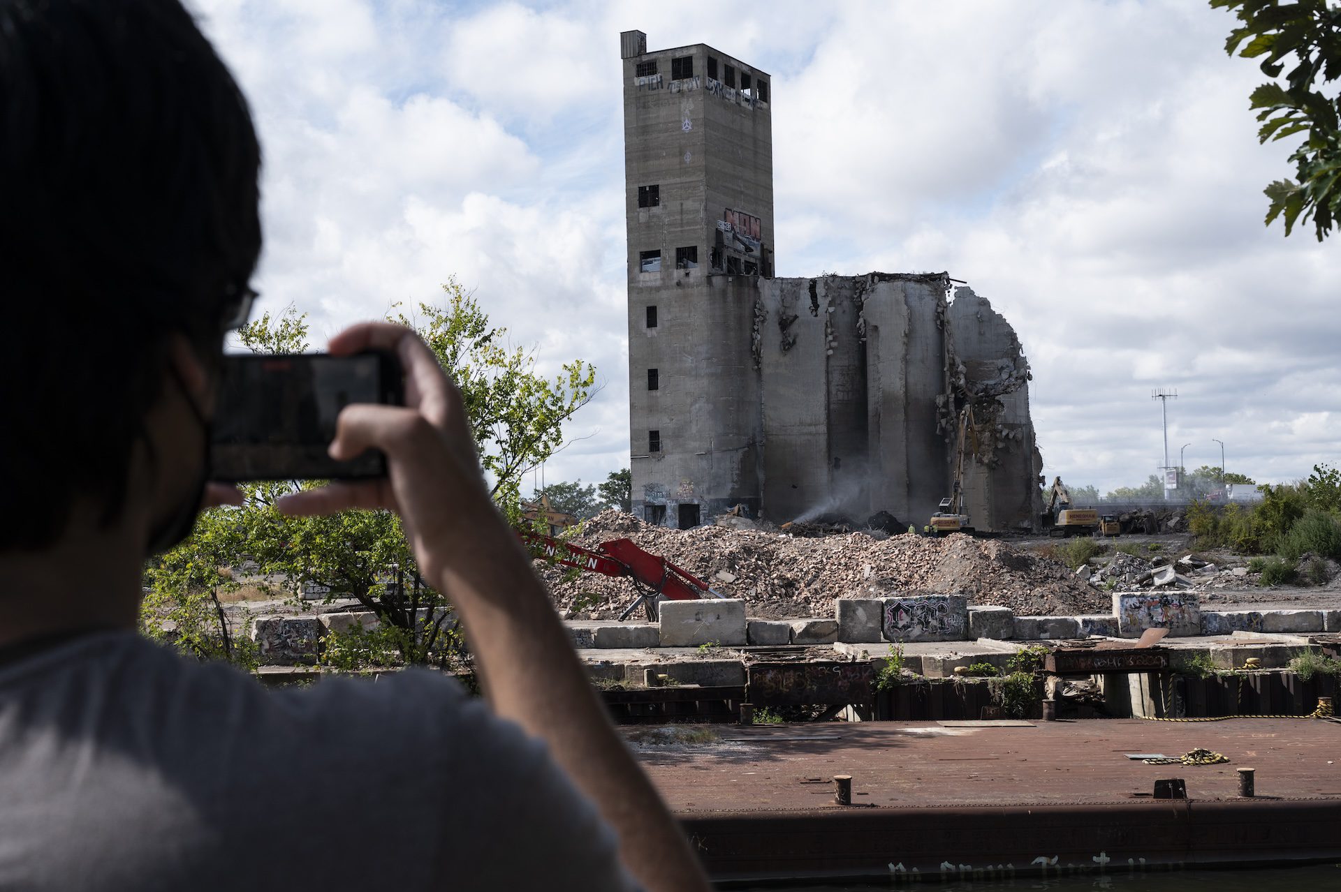 Ajay, an environmental activist, takes videos of the demolition of the Damen Silos as multiple cranes pull down the remaining silo towers, which are surrounded by rubble.