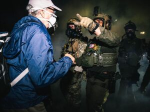 Federal agents wearing gas masks and fatigues walk through tear gas pointing the direction they are commanding protestors to go.
