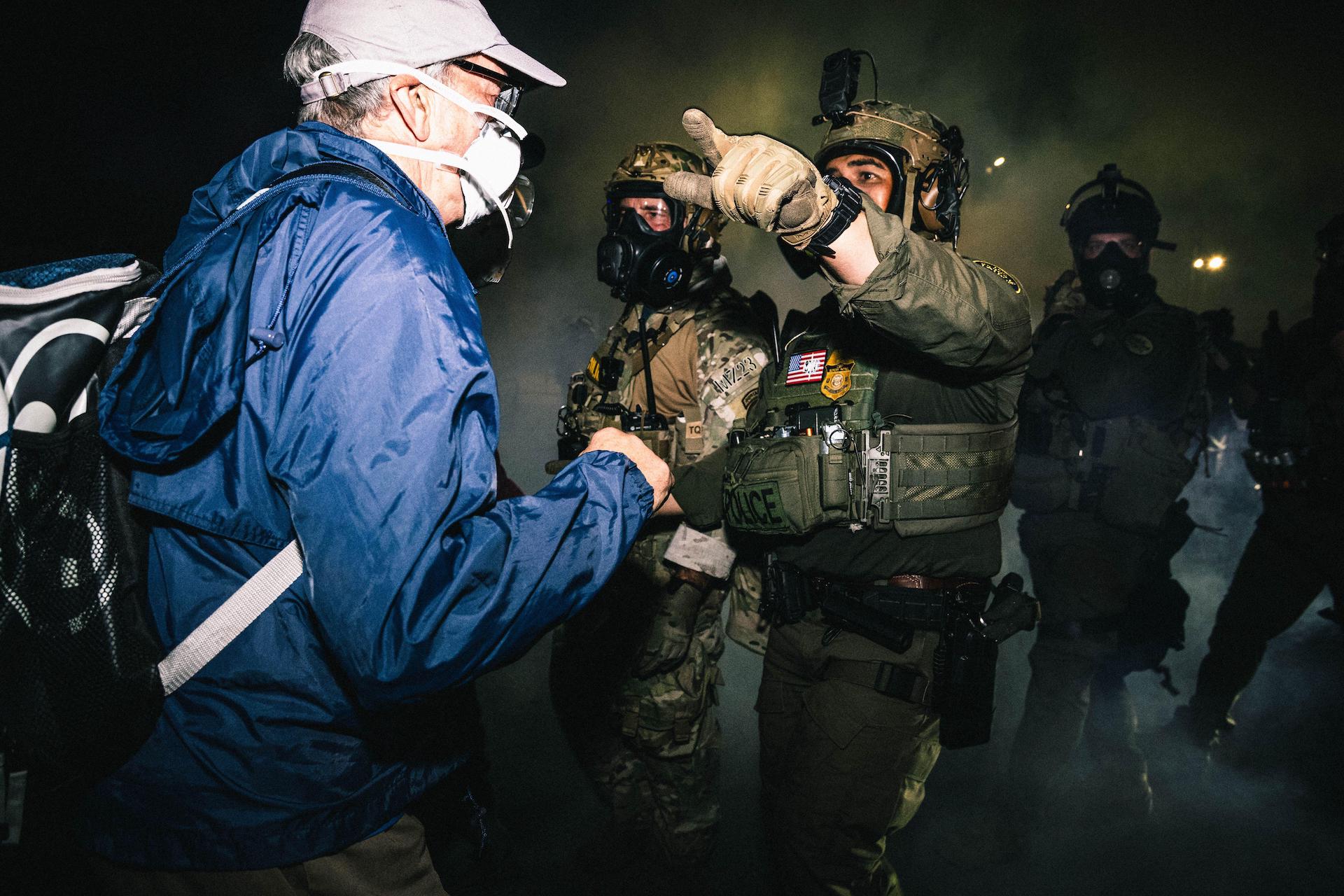 Agentes federales con máscaras antigás y uniformes de faena caminan entre gases lacrimógenos señalando la dirección que ordenan a los manifestantes.