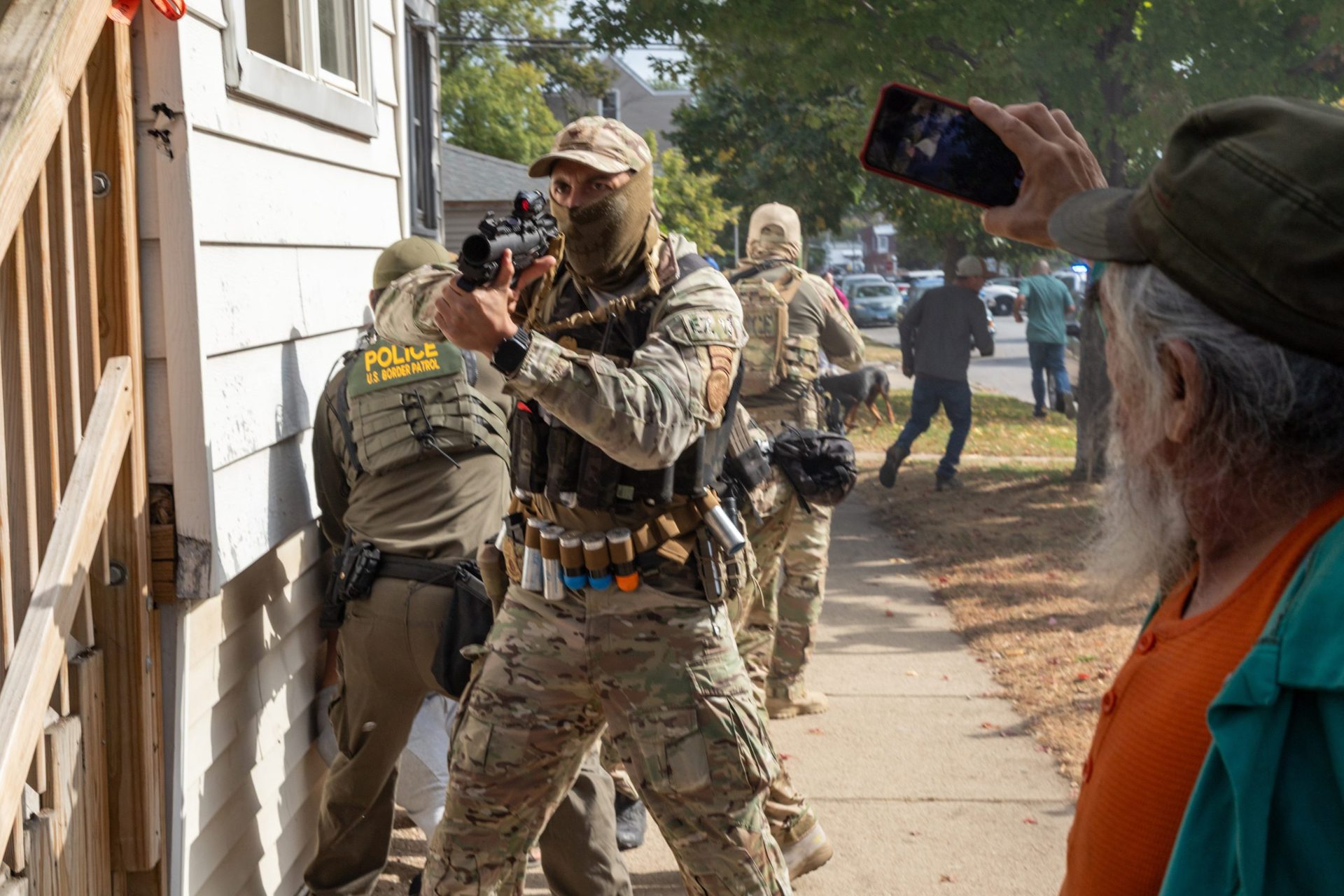 A masked federal agent points a weapon at residents on the East Side of Chicago. One elderly man is seeing filming with his phone.