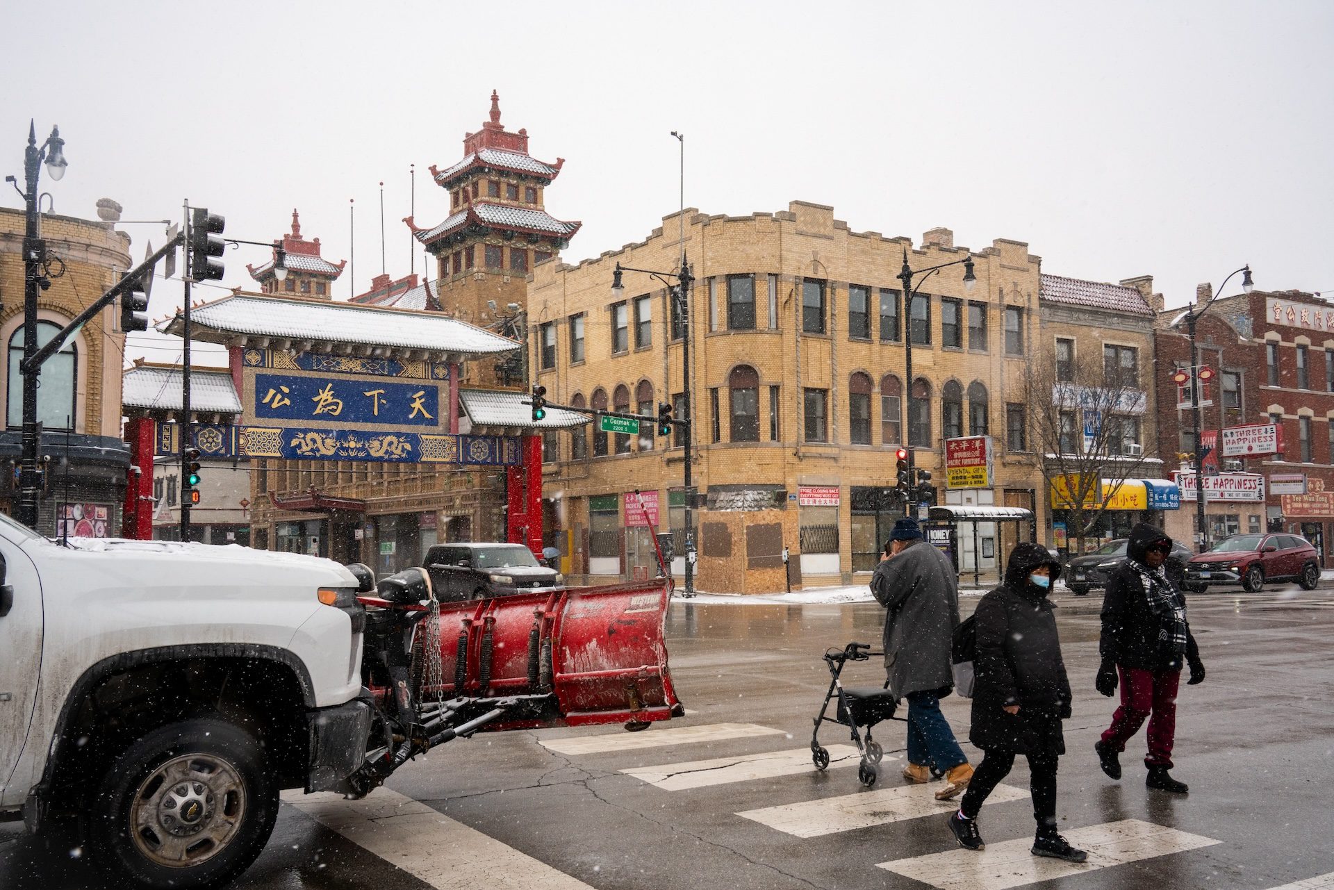 With snow falling, people cross the busy Chinatown intersection of Cermak Road and Wentworth Avenue on Jan. 30, 2026. Chicago's Chinatown Gateway is seen in the near distance.