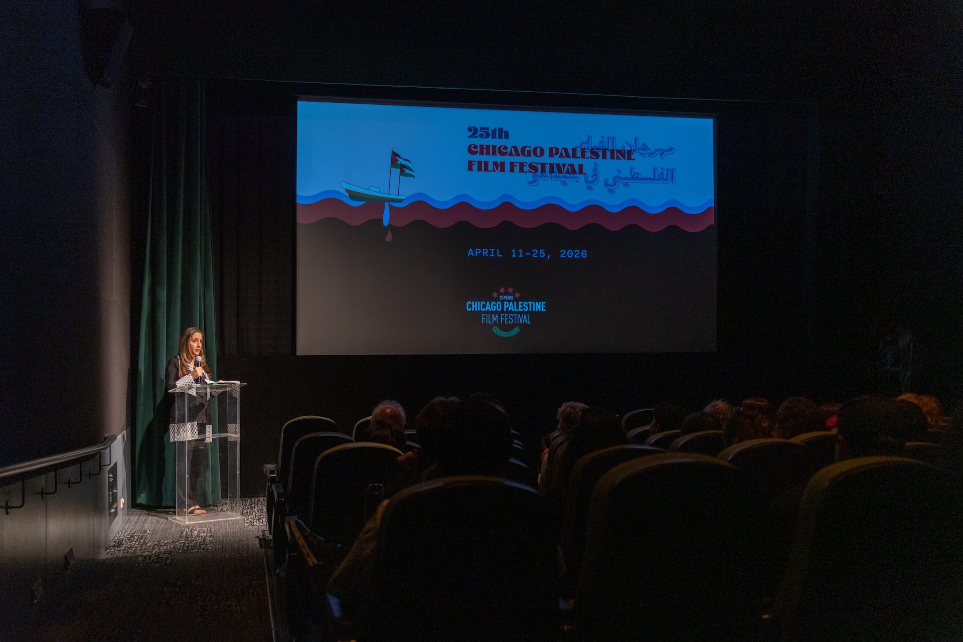 A spotlight illuminates Nina Shoman-Dajani, Executive Director of the Chicago Palestine Film Festival, as she introduces a film at the front of a dark theater at the Gene Siskel Film Center.