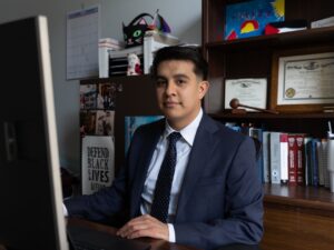 Cook County public defender Cruz Rodriguez sits at his desk in front of his computer. A gavel and degrees sit on the shelves behind him.