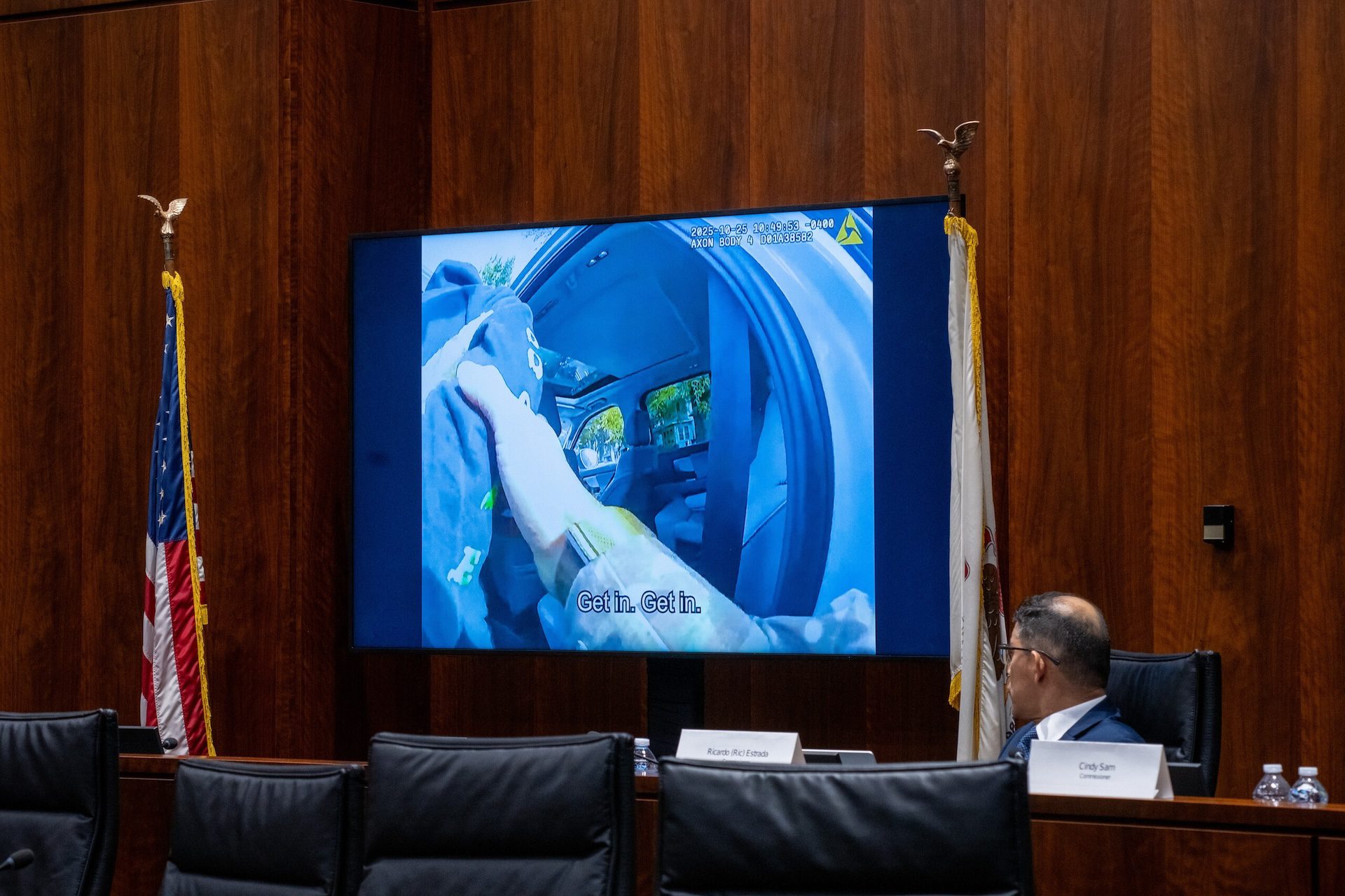 A large TV in a hearing shows body-cam footage of ICE officers interacting with protesters. Commissioner Ricardo Estrada of the Illinois Accountability Committee looks on.