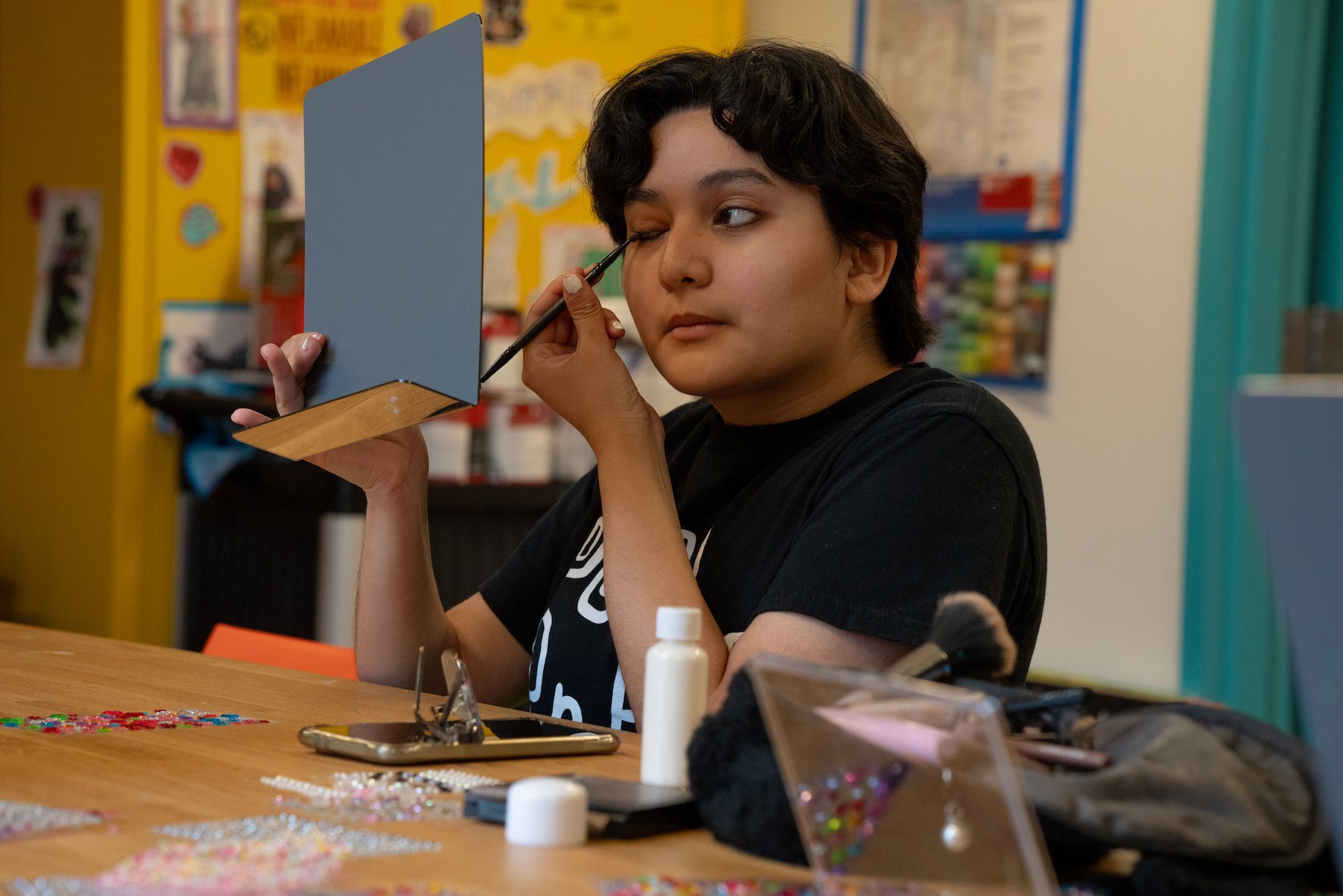 A queer prom event attendee looks in a mirror as they fix their makeup at the face gem activity table.