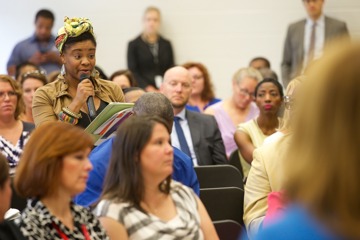 A participant stands up among a large crowd while speaking during a public education meeting.