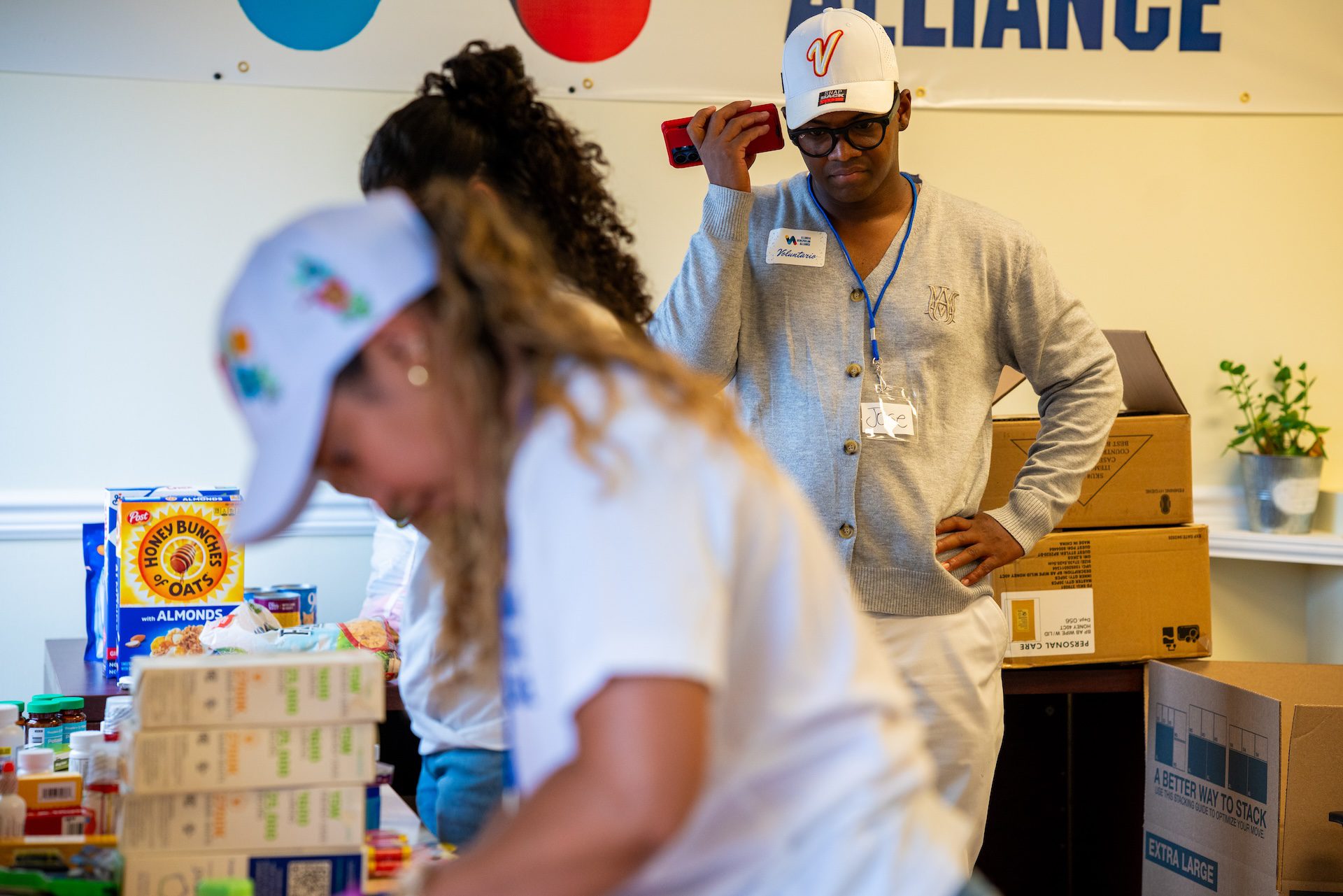José Corro sostiene su teléfono para llamar a un familiar en Venezuela mientras otros dos voluntarios empacan donaciones de alimentos en caja y artículos de primeros auxilios en la Alianza Venezolana de Illinois.