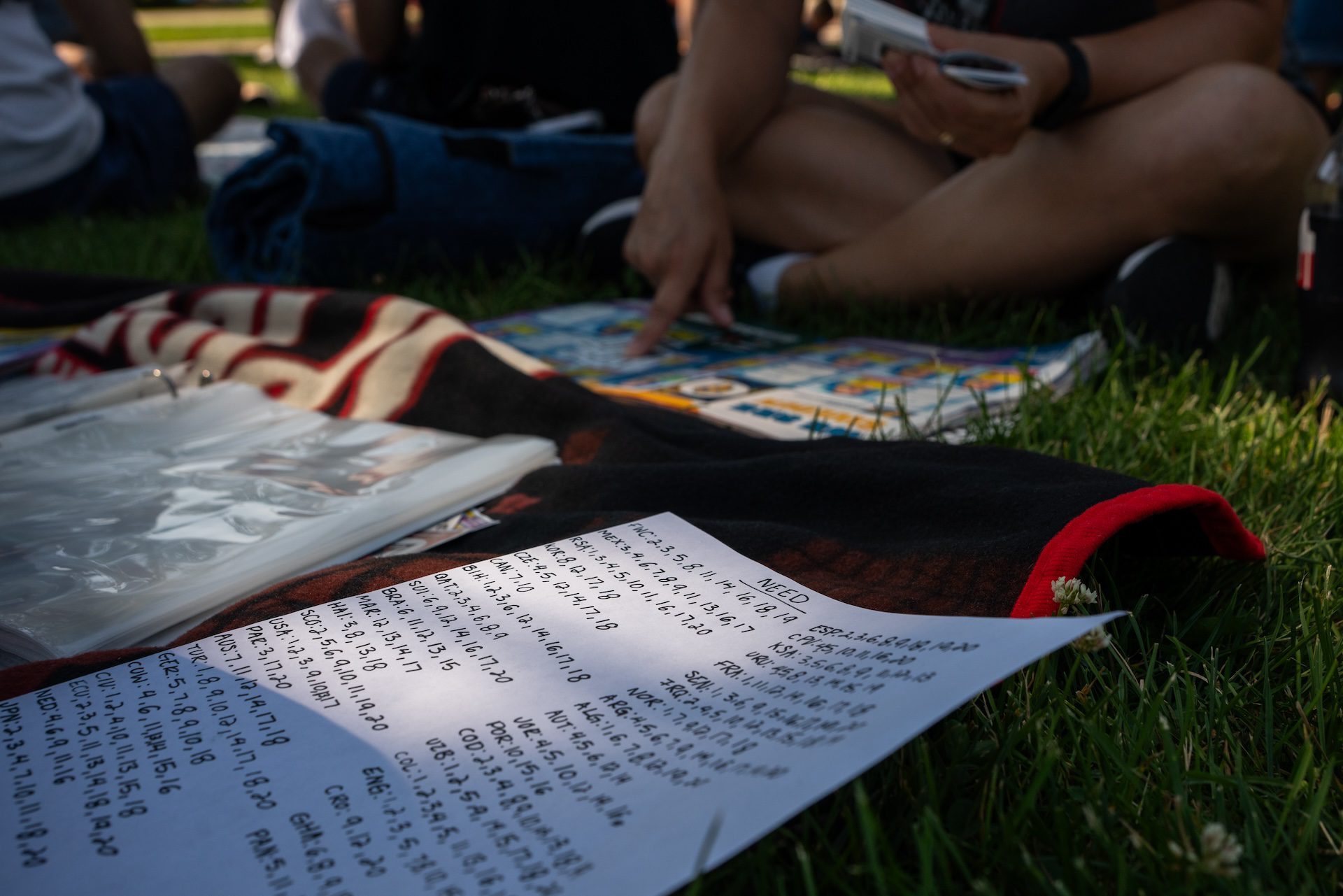 A handwritten list of card numbers is laid on the grass that shows what stickers a Panini sticker collector is still in need of.
