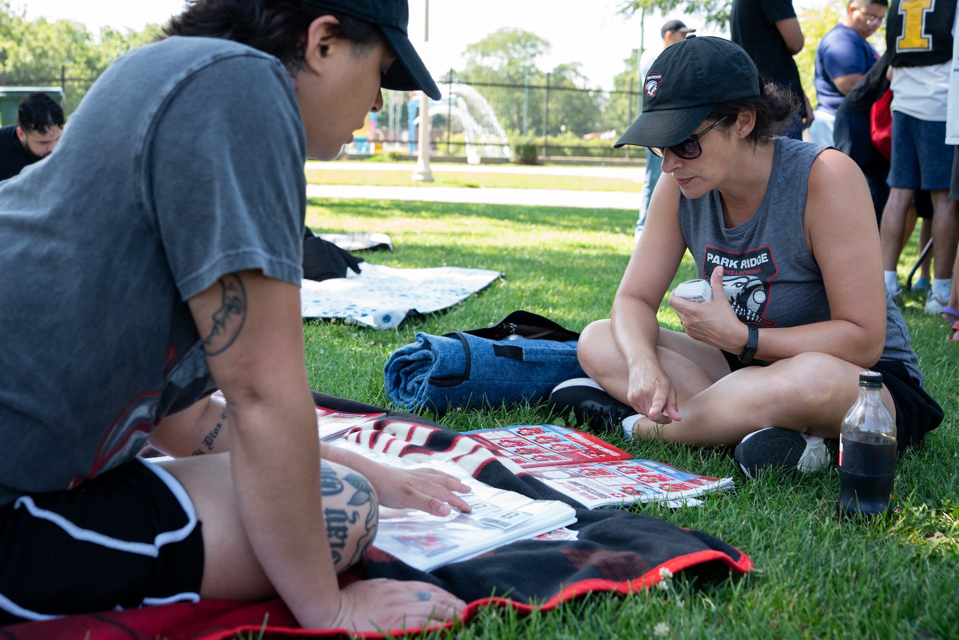 Ana Magnuson (right) sits cross legged on the grass as she trades Panini stickers with Chantal Esparza (left), seated on a blanket, during a swap meet up in Portage Park on July 12, 2026.