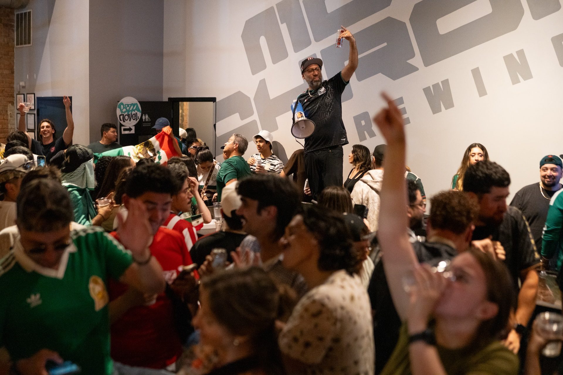 Monochrome Brewing owner Enrique Rivera stands on a ladder as he celebrates with a large group of watch party attendees after Mexico won against South Korea during the World Cup match watch party.