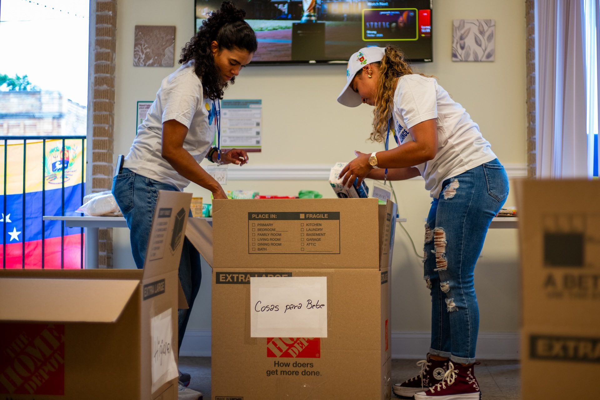 Dos voluntarios empacan donaciones de alimentos secos en una caja, artículos de primeros auxilios y productos de higiene personal en la sede de la Alianza Venezolana.