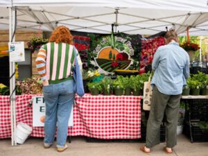 Dos clientes de espaldas buscan productos en Los Rodriguez Farms en el mercado de agricultores de Uptown el 13 de mayo de 2026.