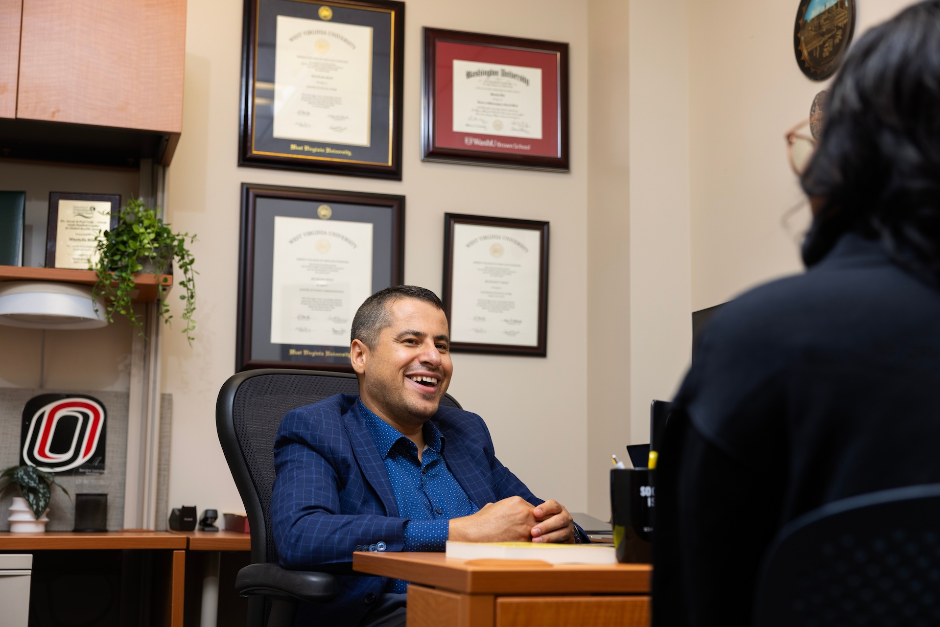 Mustafa Rfat smiles, seated in his office at the University of Nebraska at Omaha’s Grace Abbott School of Social Work with degrees and certificated framed on the wall. Rfat meets with Brittany Jones (partially pictured) who is currently working toward a Master in Social Work degree with a minor in victimology and victim services on Tuesday, Sept. 22, 2026, in Omaha.