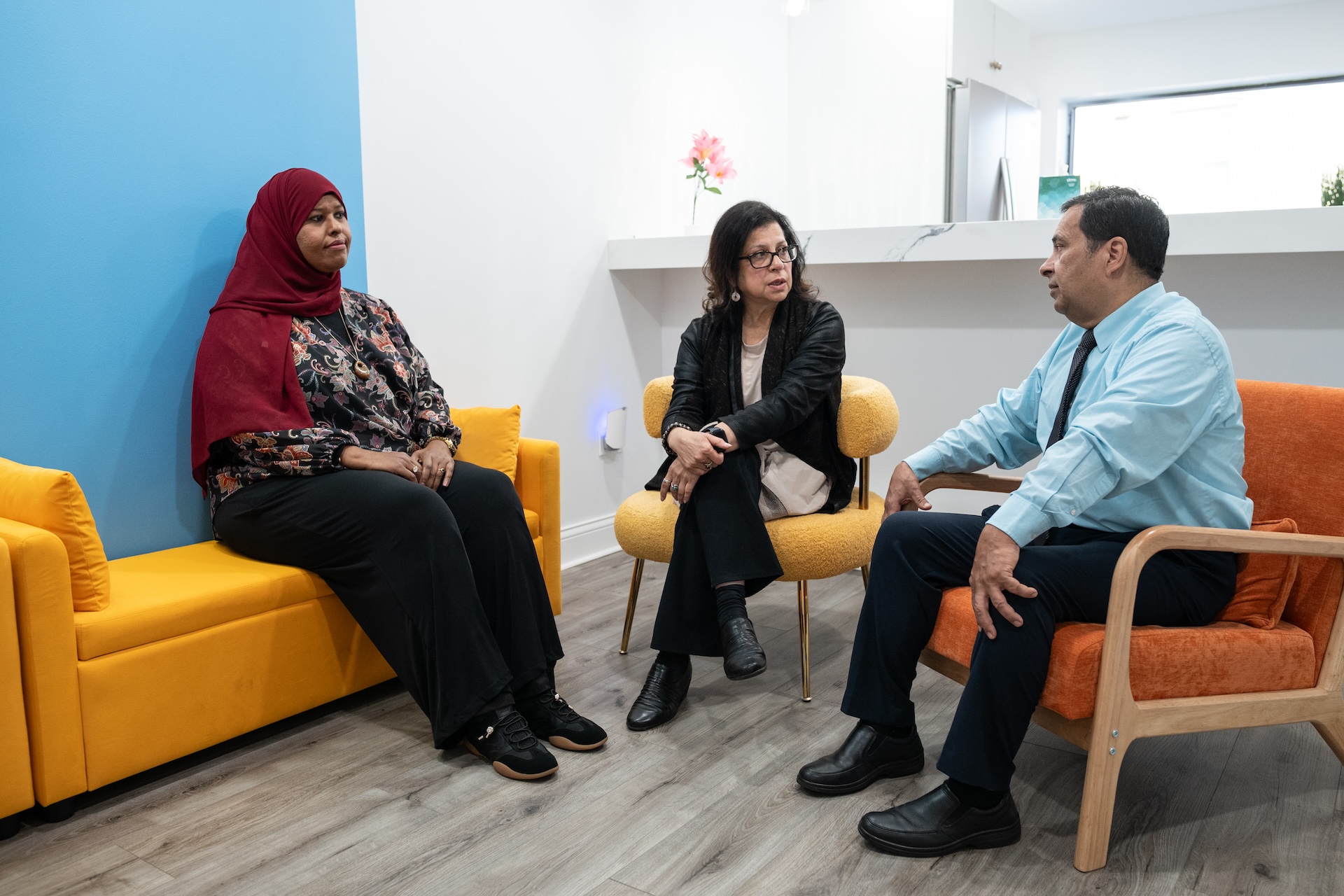 In a break room with orange and yellow cushioned furniture, Rooshey Hasnain, a clinical associate professor in the department of Disability and Human Development Health and Rehabilitation at UIC, talks about potential collaborations with Nana Ahmed, founder and CEO of Agents of Hope (L), and Francisco Alvarado, assistant deputy director of operations at the Illinois Department of Human Services (R), at Agents of Hope in West Ridge, Sept. 27, 2026. Hasnain, Ahmed and Alvarado worked together on several grant-funded projects serving refugees with disabilities, including PRIDE (Partners with Refugees in Illinois Disability Employment).
