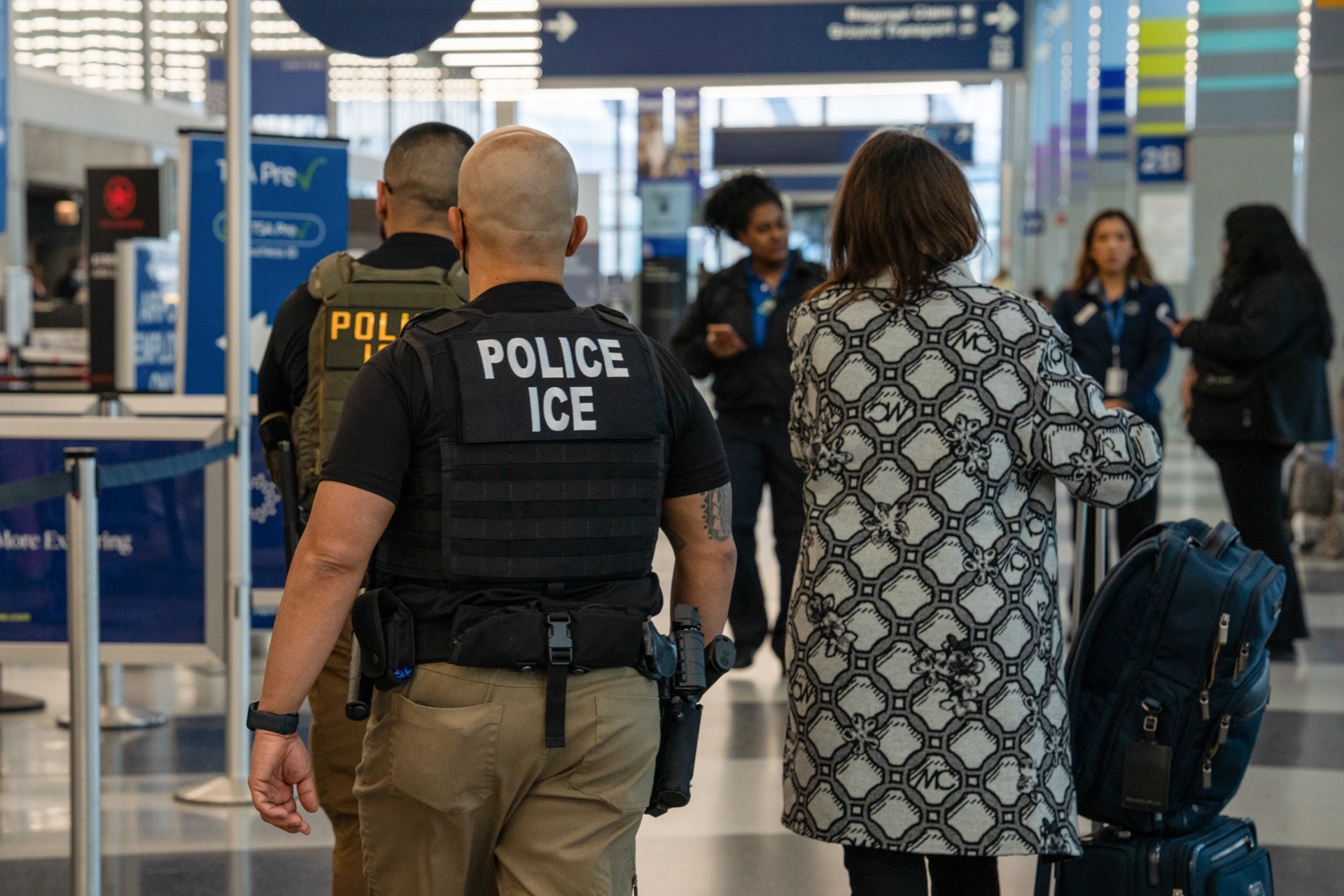 Two ICE agents wearing body armor vests walk alongside travelers at O'Hare International airport.